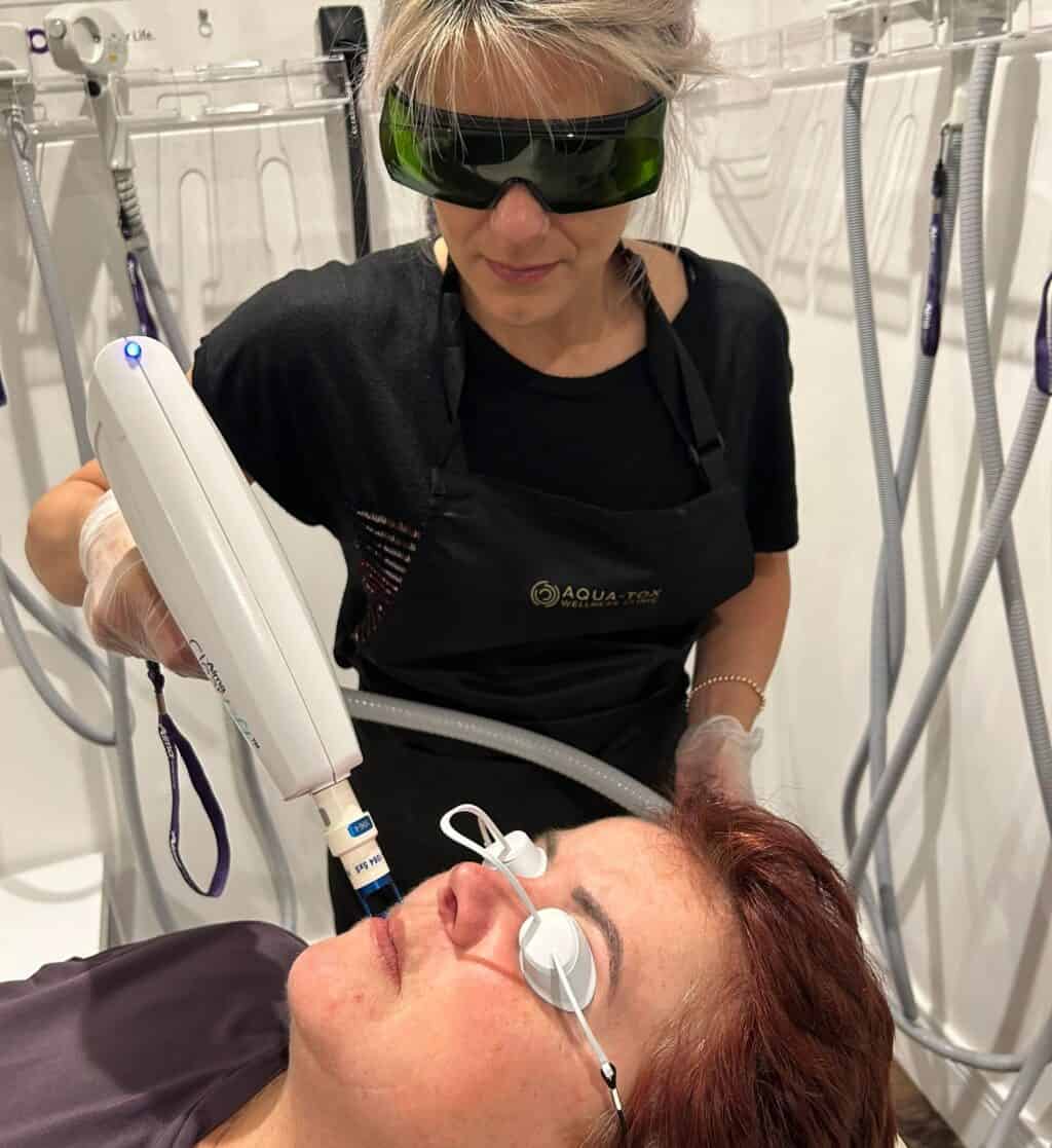 A female therapist is holding a laser skin device to the face of a female laying on a beauty couch. The female on the bed is wearing small protective goggles.