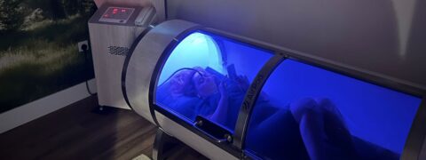 A female is inside a hyperbaric oxygen chamber lit with blue light. She is looking at a mobile phone and lying on her back
