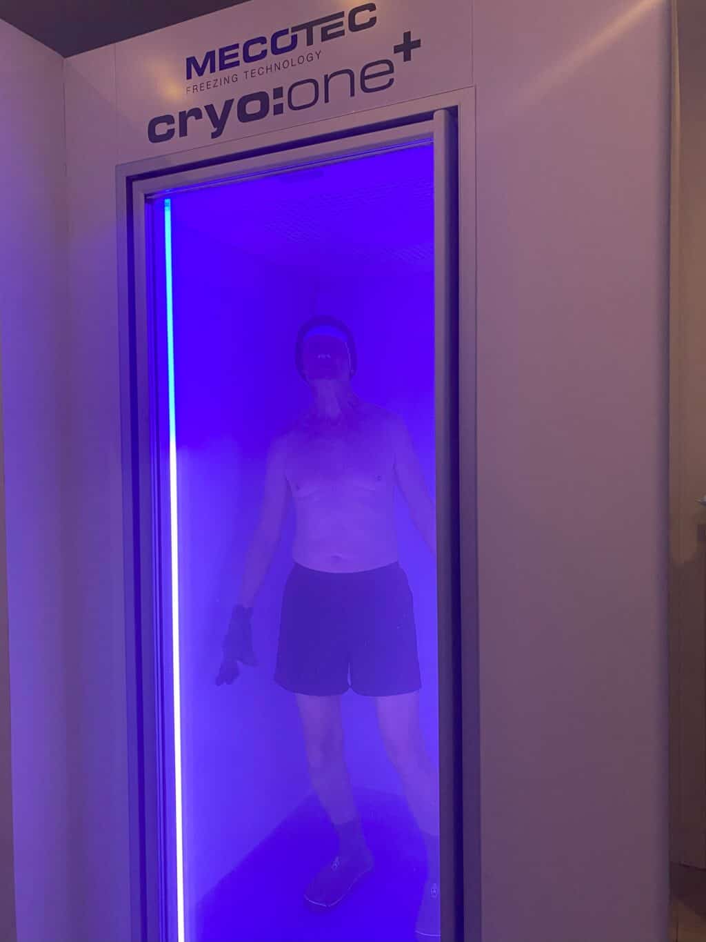 a 71 year old man inside a mecotec cryo one cryotherapy chamber, he is shirtless and the chamber is lit with blue light