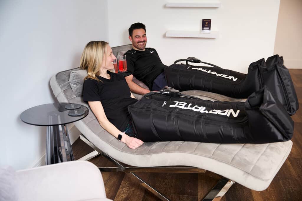 a man and a woman are laying on grey chaise lounges wearing norma tec leg compression boots