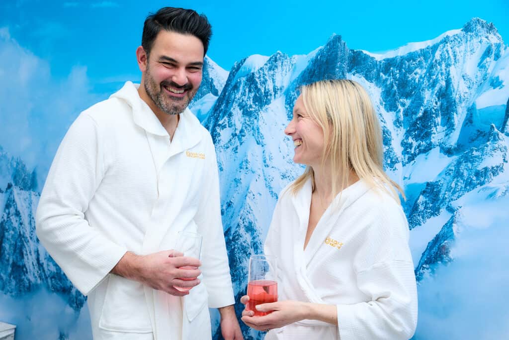 a man and a woman are wearing white robes and holding drinks against a backdrop wall of a snowy mountain