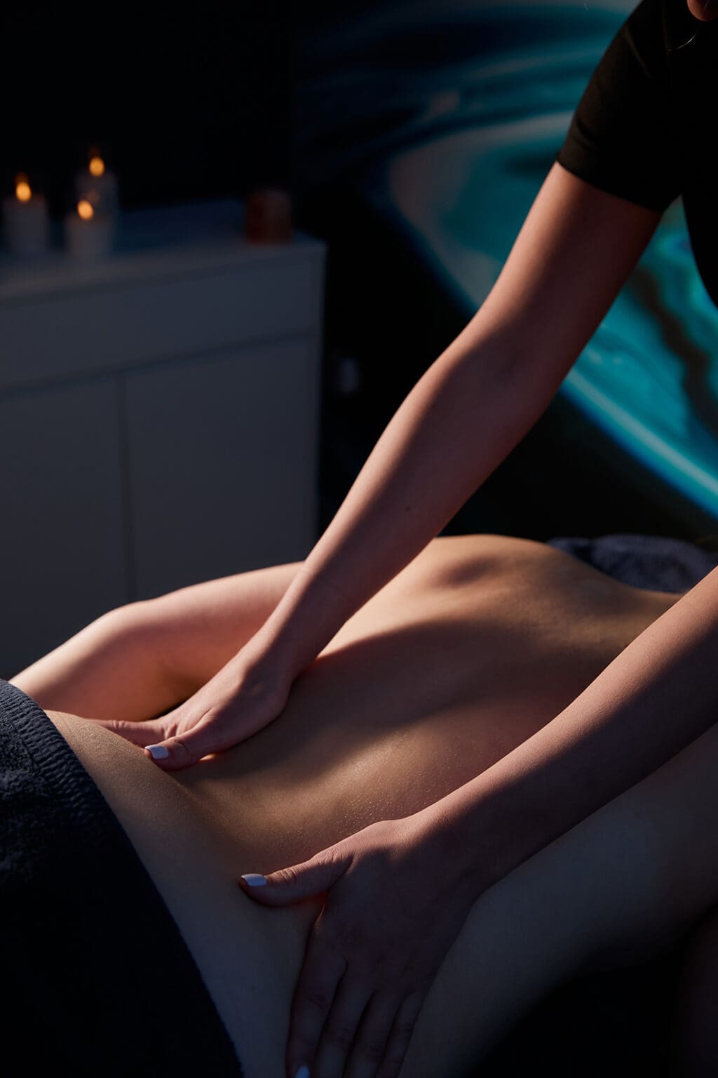 a massage therapists hands can be seen giving a back massage in a darkened room, there are candles in the background