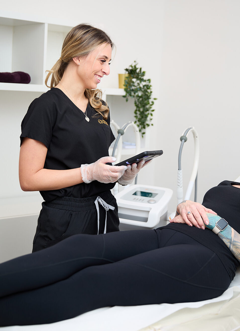 a female therapist wearing black scrubs is holding an ipad and talking to a client on a treatment bed, there is a radiofrequency facial device in the background