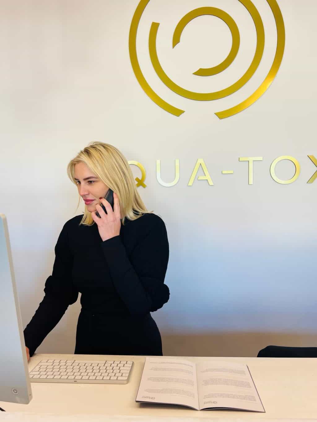 a blonde haired female standing at a reception desk on the phone with an aqua-tox logo before