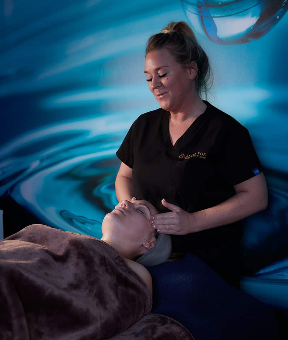 a female therapist wearing black scrubs performs a facial on a female covered with a blanket on a treatment couch in a darkened room in Hertford